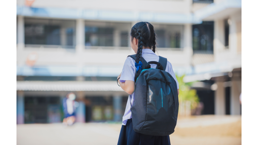 A young girl walks towards a school building with a blue pack on her back