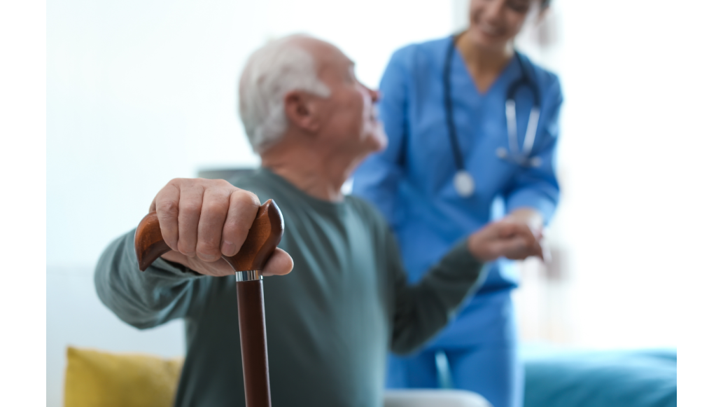 An elderly man holding onto a cane is assisted by a clinician