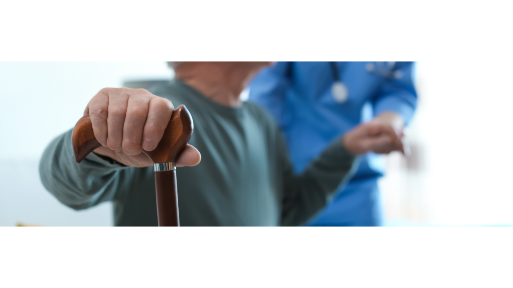 An elderly man holding onto a cane is assisted by a clinician