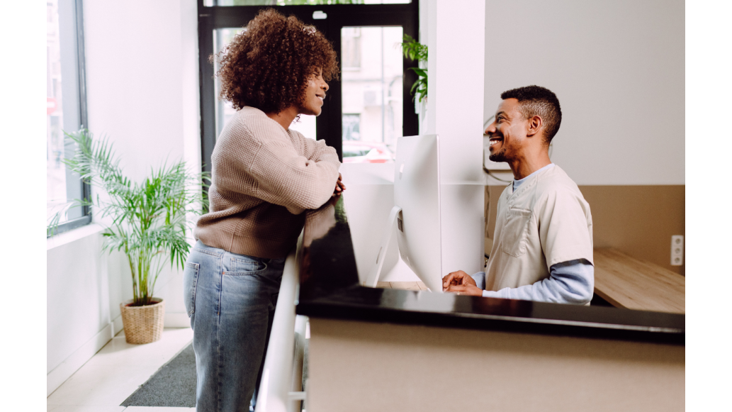 A woman stands at a doctor's office reception desk speaking with the male receptionist