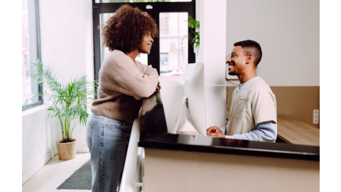 A woman stands at a doctor's office reception desk speaking with the male receptionist