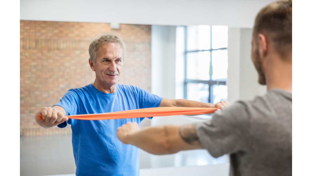 An older man stretched a resistance band while facing a trainer