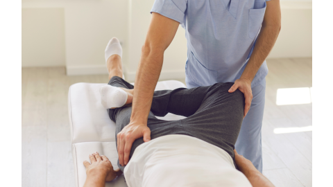 A male clinician assists a man lying on a treatment table with a pelvic "figure four" stretch