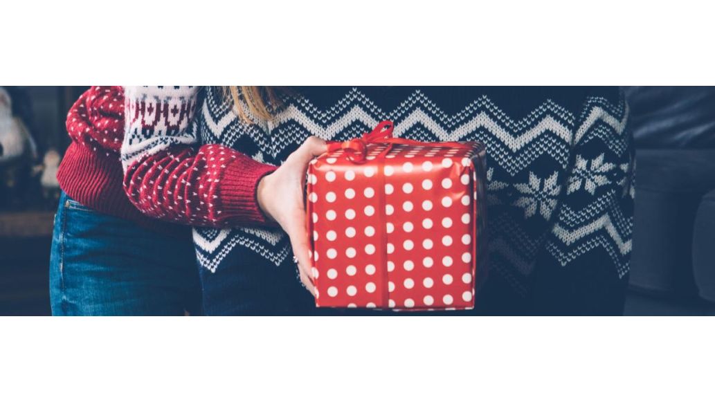woman with present in hand hugs man from behind, presenting it to him with one hand covering his eyes