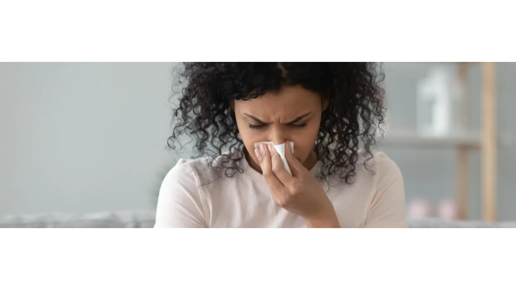 Woman sitting at laptop while holding tissue to her nose as if she's sneezing.