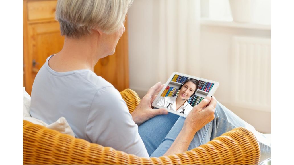 Person sitting on couch, holding tablet with doctor on screen conducting telehealth visit