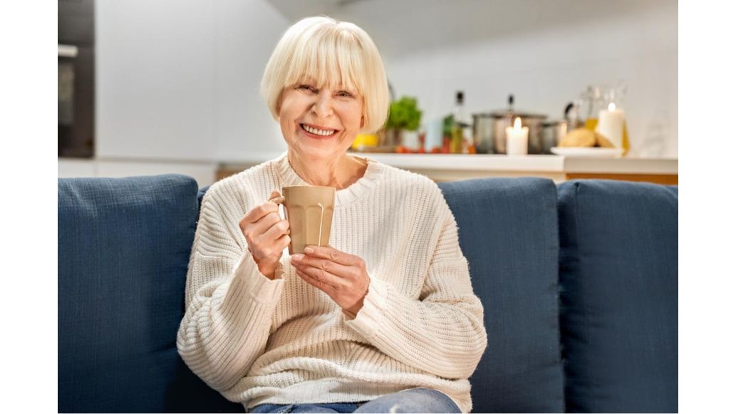Older woman seated on couch, holding mug and smiling.