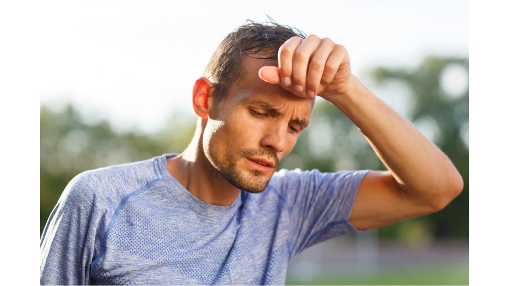 Man with hand at his head, sweating & slightly leaned over as if he's exhausted from working out outside in the heat.