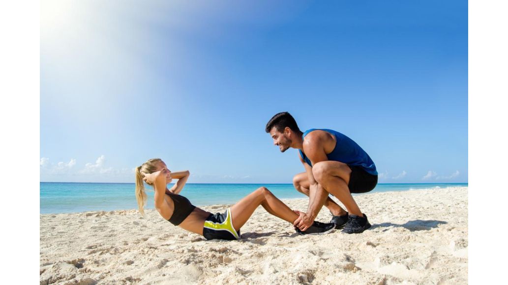 Woman doing sit up exercise on the beach while a partner holds her feet in place on the sand.