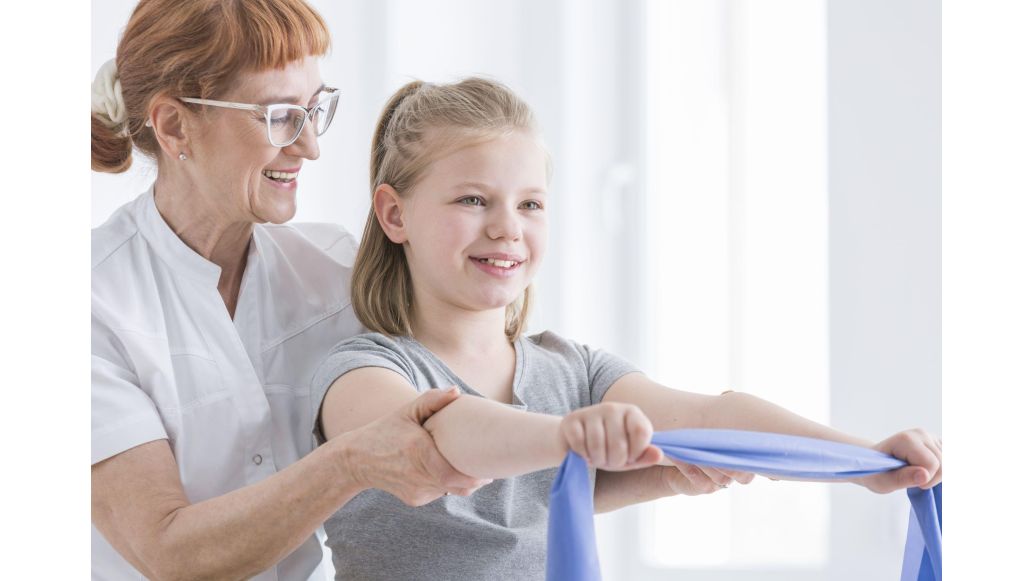 Young girl smiling, holding a light blue resistance band with arms outstretched and with assistance from physiotherapist.